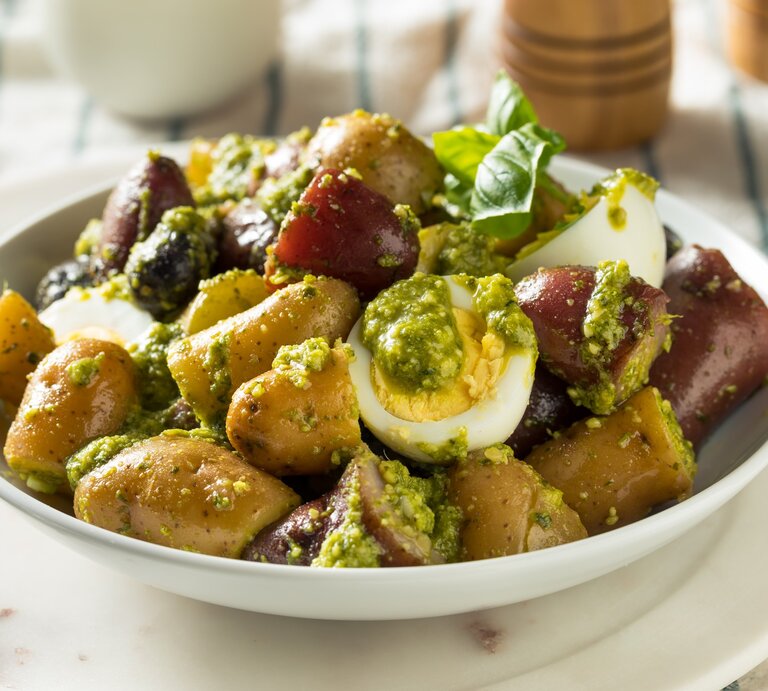 Kartoffelsalat mit grünem Pesto, Oliven und Ei in weißer Salatschüssel auf weißem Anrichteteller auf blau-weißem Tuch. Daneben Basilikumblätter. Darüber Pfeffermühle aus Holz. Frontalaufnahme. | © Getty Images / bhofack2