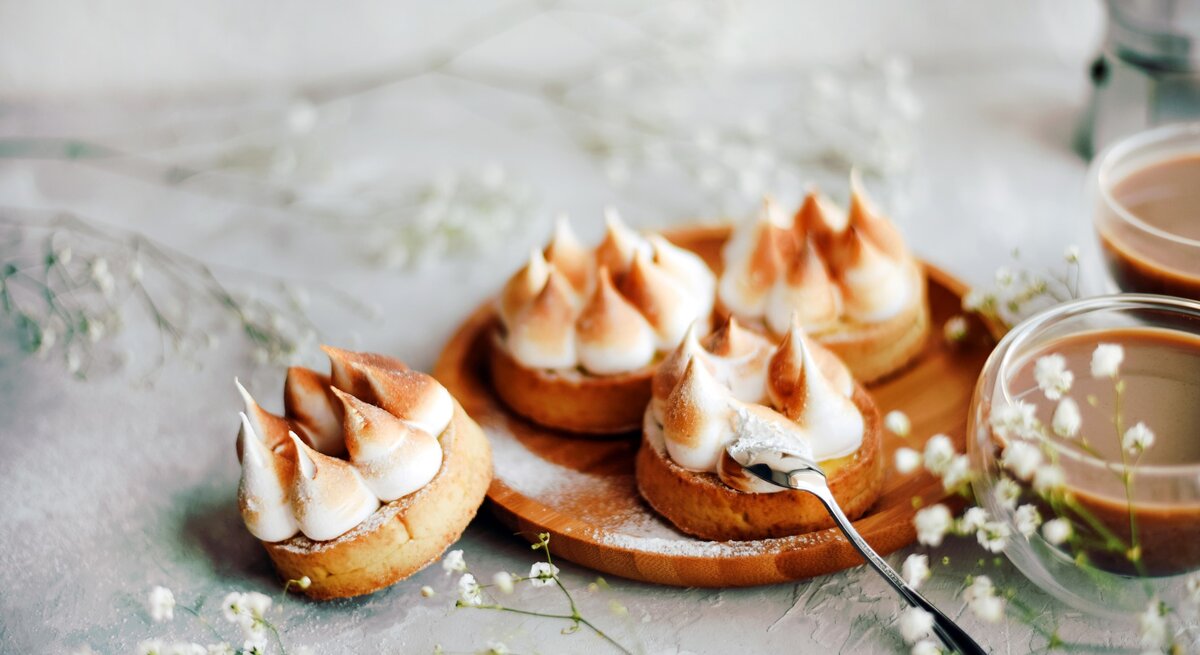 Vier Mini-Törtchen mit Wintercreme und Baiser aus Plätzchenteig auf Holzbrett. Daneben zwei Gläser mit Kaffee oder Kakao. Im Bild Blüten als Deko verteilt. Frontalaufnahme.
