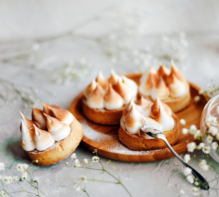 Vier Mini-Törtchen mit Wintercreme und Baiser aus Plätzchenteig auf Holzbrett. Daneben zwei Gläser mit Kaffee oder Kakao. Im Bild Blüten als Deko verteilt. Frontalaufnahme. | © Getty Images / Maryna Terletska