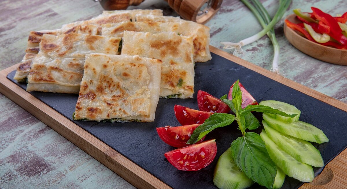 Gözleme mit Spinat-Schafskäsefüllung auf Brett mit Salat. Daneben Lauch und Gewürzmühlen aus Holz. In eienr Schale Paprikastreifen. Draufsicht.