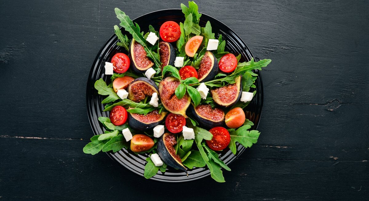 Vogelperspektive: Ein Feigen-Salat mit Rucola und Feta auf einem dunklen Teller, der auf einem dunken Untergrund steht.