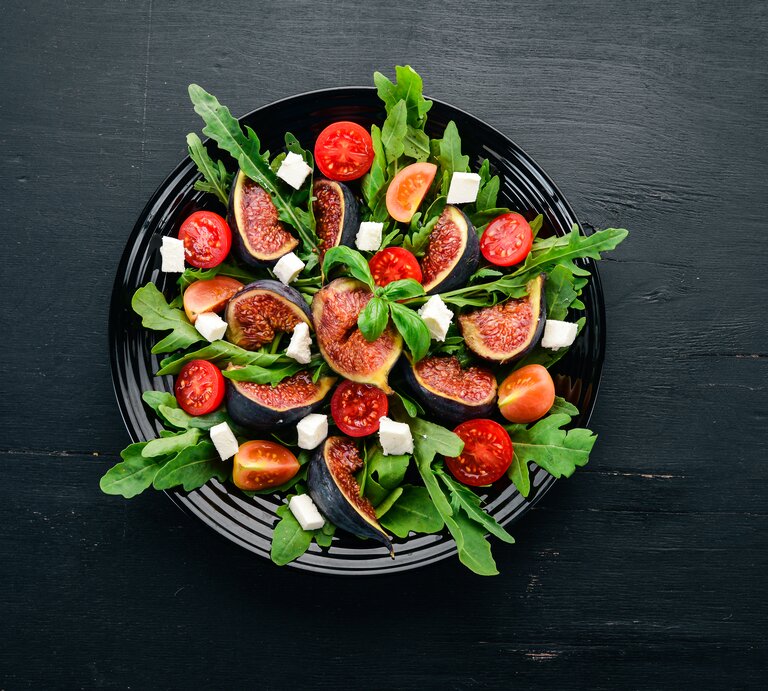Vogelperspektive: Ein Feigen-Salat mit Rucola und Feta auf einem dunklen Teller, der auf einem dunken Untergrund steht. | © Shutterstock / YARUNIV Studio
