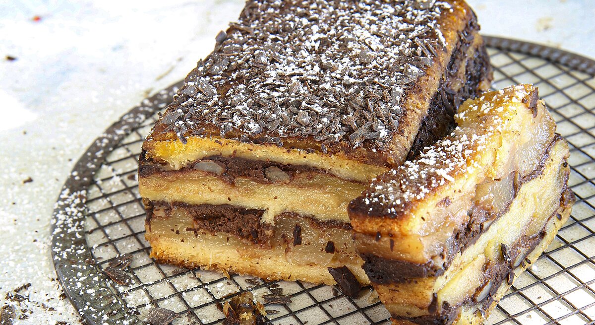 Croque Cake mit Apfel-Birnen-Schokolade, bestehend aus mehreren Schichten Kastenkuchen, getoppt mit Schichten aus Birne und Schokoladencreme und bestreut mit Puderzucker und Schokostreuseln. 