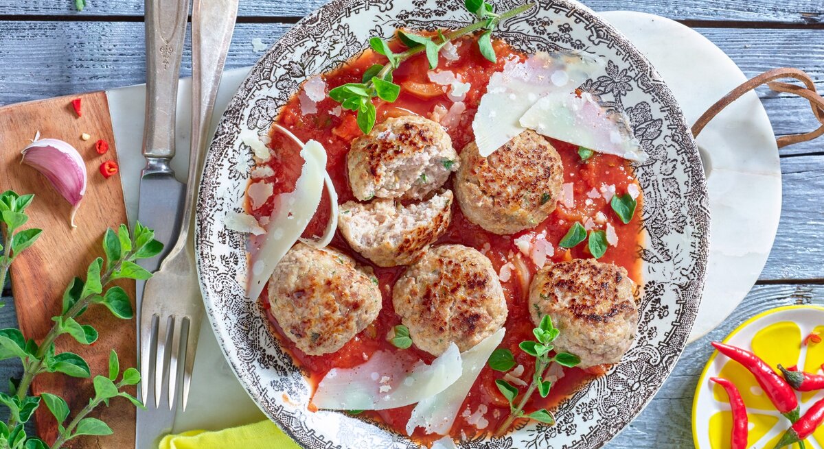 Hackbällchen mit Ricotta und Oregano, serviert auf einem Sud aus Tomaten, garniert mit frischen Kräutern und groben Parmesanspänen auf einem traditionell italienisch dekorierten Teller, platziert auf einem hellen Holztisch. 