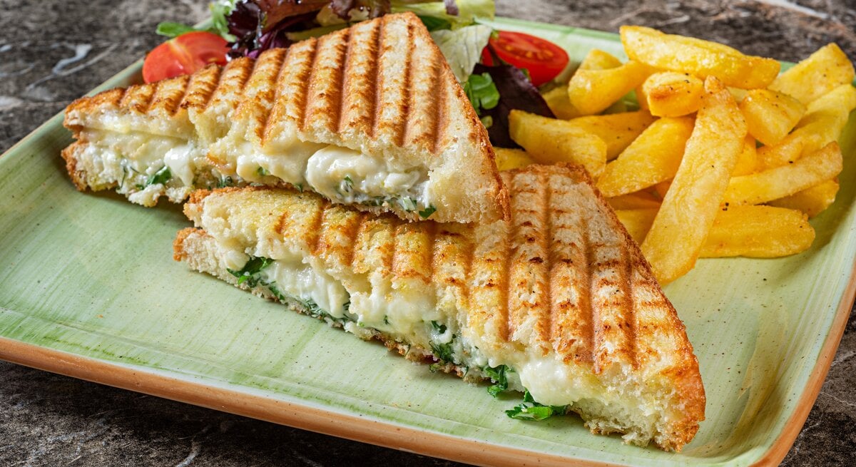 Auf einem eckigen Teller liegen zwei Panini mit Brokkoli und Käse gefüllt, außerdem Salat und Fritten.