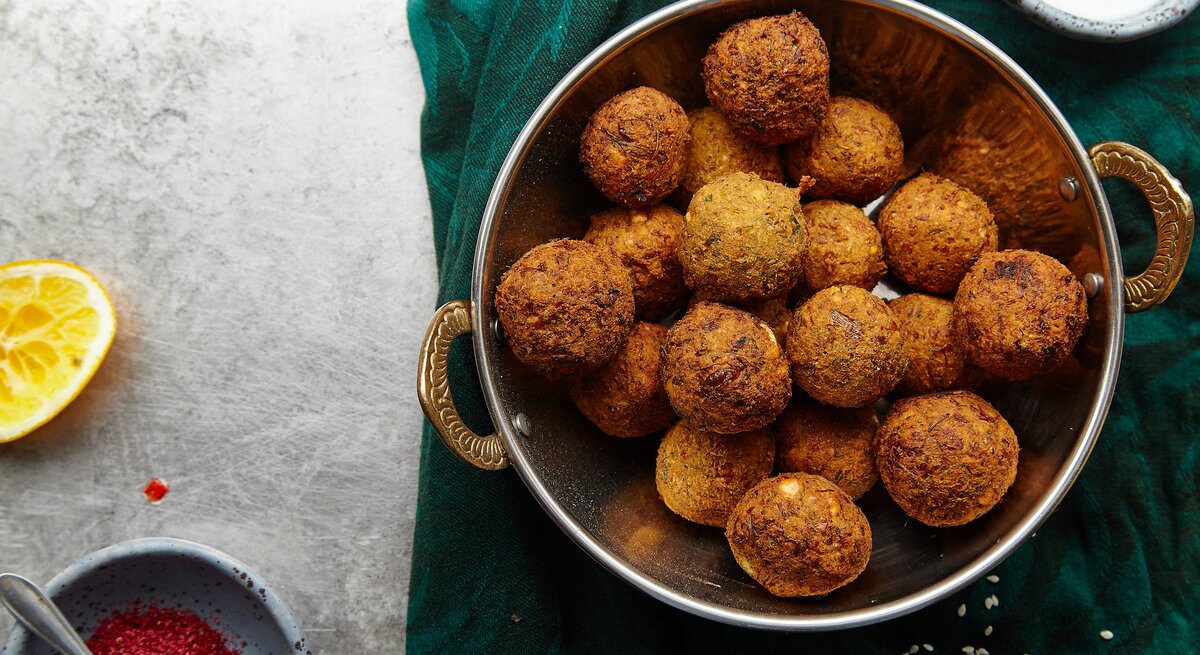 Eine silberne Schale gefüllt mit Maronen-Falafel mit Cranberrys neben einer Schüssel mit roten Gewürz, einer ausgepressten Zitronenhälfte und einer Schüssel mit einem Joghurt-Dip, vor einem dunkel grün und grauen Hintergrund, von oben. 