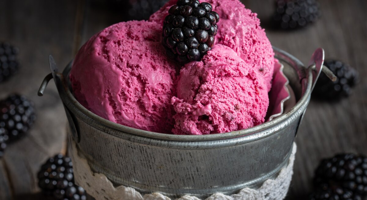 Ein kleiner Metalleimer mit 3 Kugeln Brombeer-Bananen-Eis, garniert mit einer Brombeere, dunkle Umgebung.