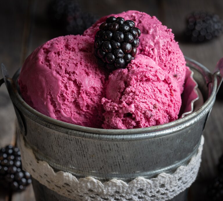 Ein kleiner Metalleimer mit 3 Kugeln Brombeer-Bananen-Eis, garniert mit einer Brombeere, dunkle Umgebung. | © Adobe Stock/ AndreaR
