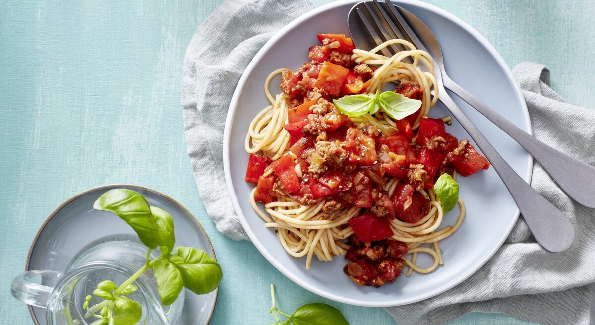 Pasta mit Hack und Paprika, garniert mit frischem Basilikum und serviert auf einem Teller auf hellblauem Tisch. 