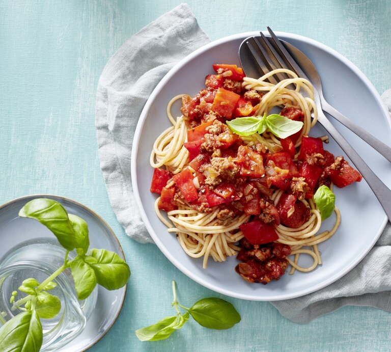 Pasta mit Hack und Paprika, garniert mit frischem Basilikum und serviert auf einem Teller auf hellblauem Tisch.  | © EAT CLUB / Isabell Triemer