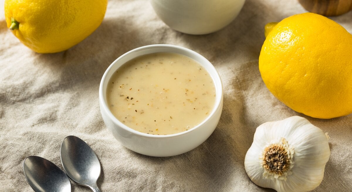 Eine Schale Zitronen-Knoblauch-Vinaigrette, zwei Zitronen, ein Knoblauch und zwei Löffeln liegen auf einem großen Leinentuch.