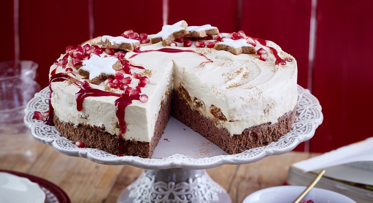 Eine angeschnittene Zimtstern-Tiramisu-Torte  mit Zimtsternen und Granatapfelkernen garniert, auf einem Kuchenständer neben einer Schüssel Granatapfelkerne vor einem roten Holzhintergrund. 