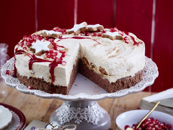 Eine angeschnittene Zimtstern-Tiramisu-Torte  mit Zimtsternen und Granatapfelkernen garniert, auf einem Kuchenständer neben einer Schüssel Granatapfelkerne vor einem roten Holzhintergrund.  | © EAT CLUB/Isabell Triemer 