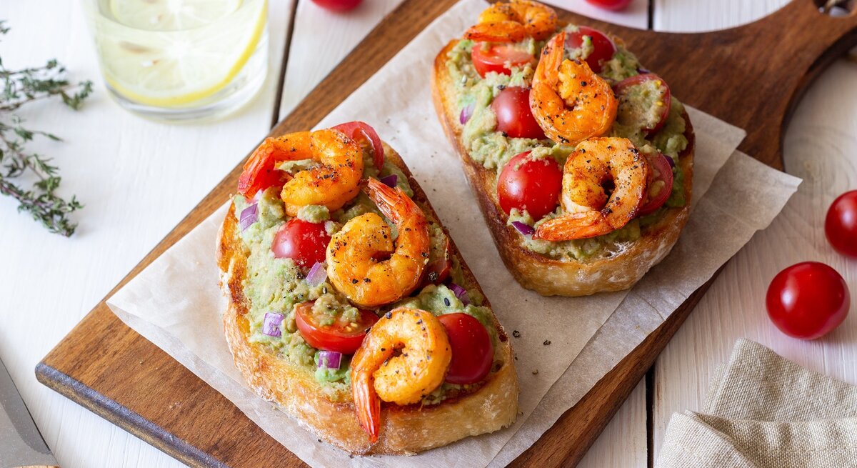 Zwei Scheiben Weißbrot auf einem Holzbrett mit Guacamole und gebratenen Garnelen.