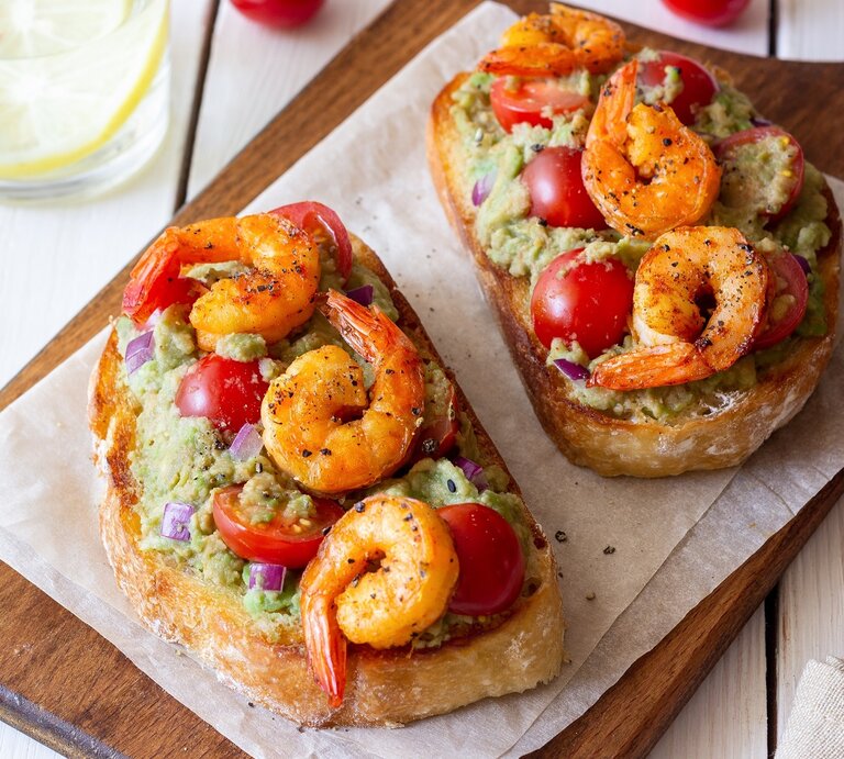 Zwei Scheiben Weißbrot auf einem Holzbrett mit Guacamole und gebratenen Garnelen. | © Shutterstock/ Vladislav Chusov