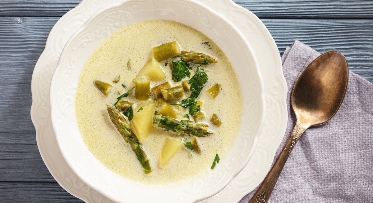 Spargel-Käse-Süppchen in verspielter Schüssel auf blauem Holz. 