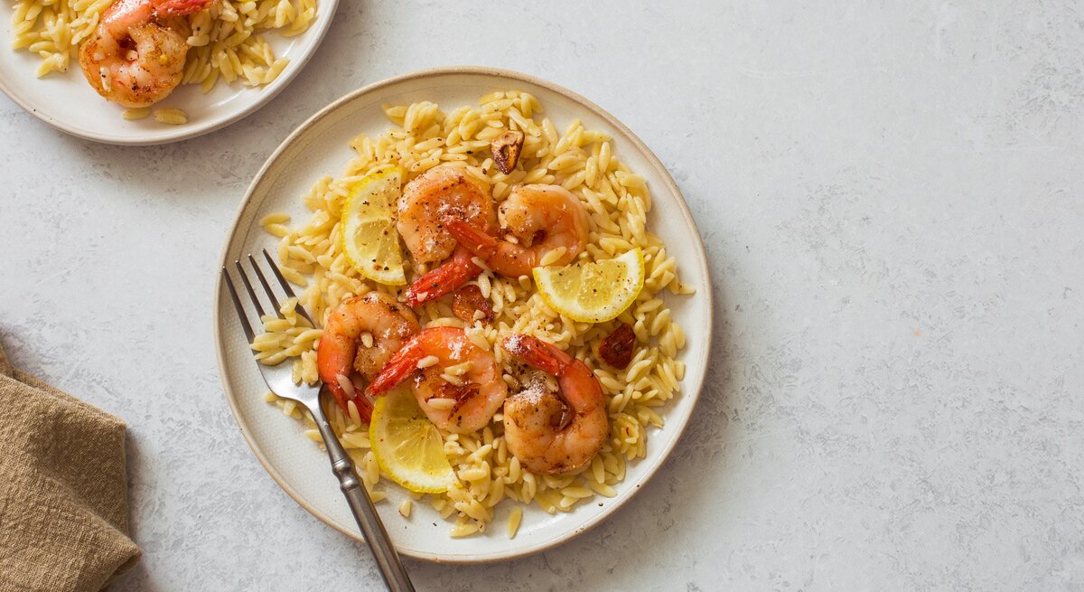 Draufsicht: Zwei Teller mit Orzo-Pasta, Zitronenscheiben und Garnelen. Auf einem Teller liegt eine Gabel.
