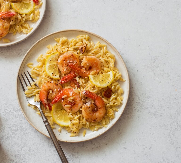 Draufsicht: Zwei Teller mit Orzo-Pasta, Zitronenscheiben und Garnelen. Auf einem Teller liegt eine Gabel. | © Shutterstock / annata78