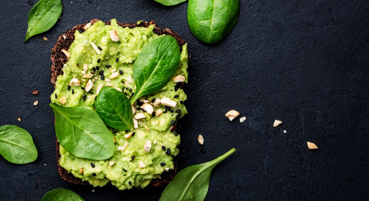 Draufsicht: Dunkler Untergund, ein Avocado-Cashew-Sandwich, dass mit schwarzen Sesamkörnern garniert ist. Drumherum liegen Spinatblätter.