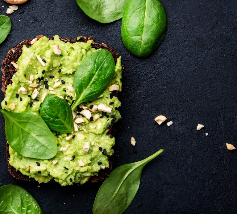 Draufsicht: Dunkler Untergund, ein Avocado-Cashew-Sandwich, dass mit schwarzen Sesamkörnern garniert ist. Drumherum liegen Spinatblätter. | © Shutterstock / 5PH