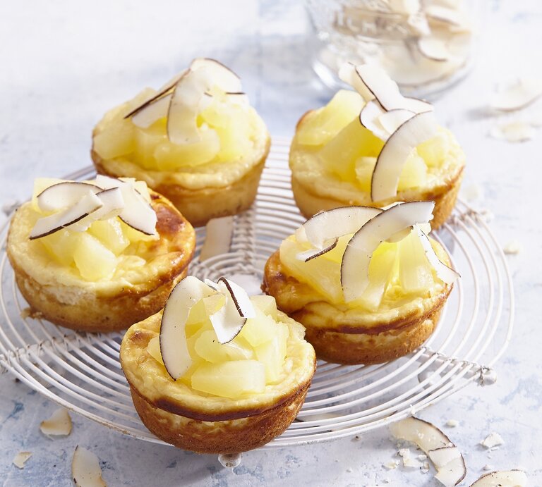 Auf einem Kuchengitter stehen mehrere Pina-Colada-Mini-Käsekuchen. Sie sind mit Kokosraspeln verfeinert.  | © StockFood / Profimedia