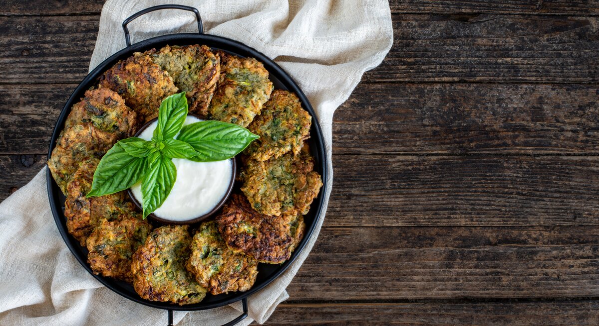 Eine Pfanne mit Brokkoli-Kartoffelpuffern, die im Kreis angeordnet sind, in der Mitte eine Schale mit Dip. Draufsicht.