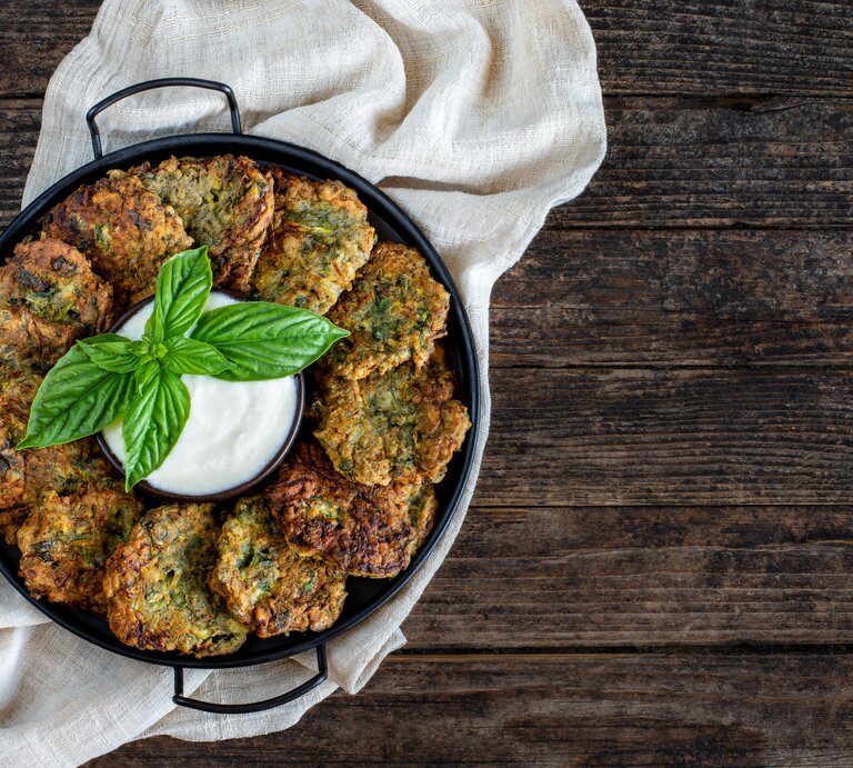 Eine Pfanne mit Brokkoli-Kartoffelpuffern, die im Kreis angeordnet sind, in der Mitte eine Schale mit Dip. Draufsicht. | © Getty Images/ Gulcin Ragiboglu