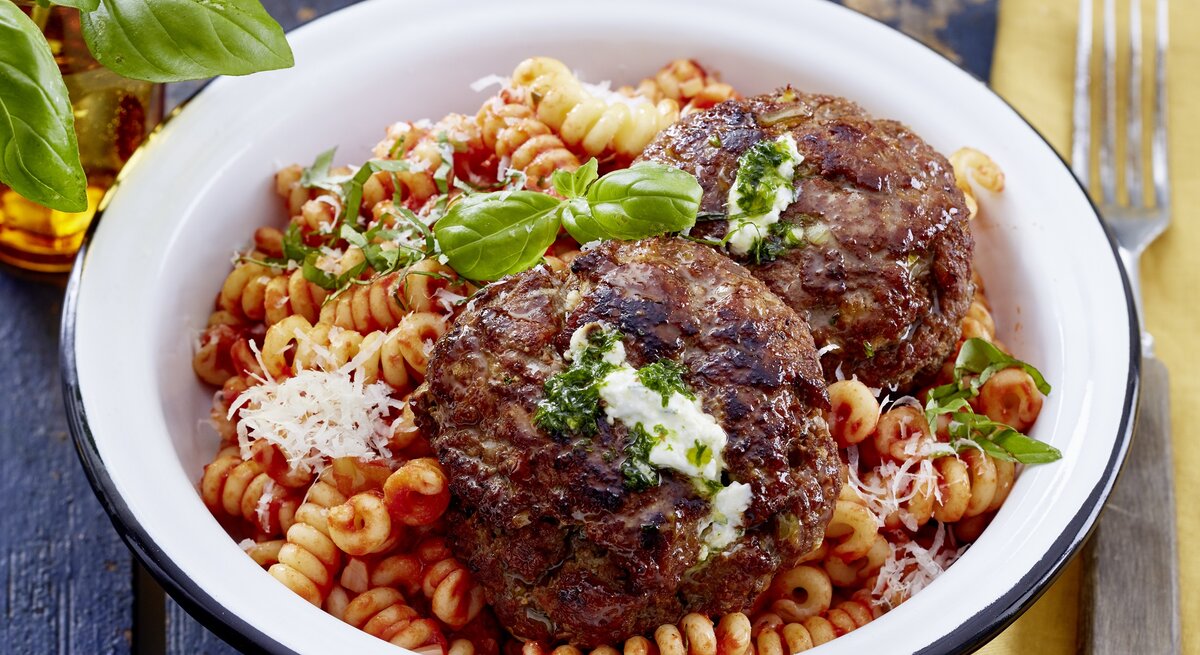 In einer Schüssel ist eine Portion Frikadellen mit Pesto-Ricotta-Füllung und Nudeln. Das Gericht ist verfeinert mit Parmesan und Basilikum. 