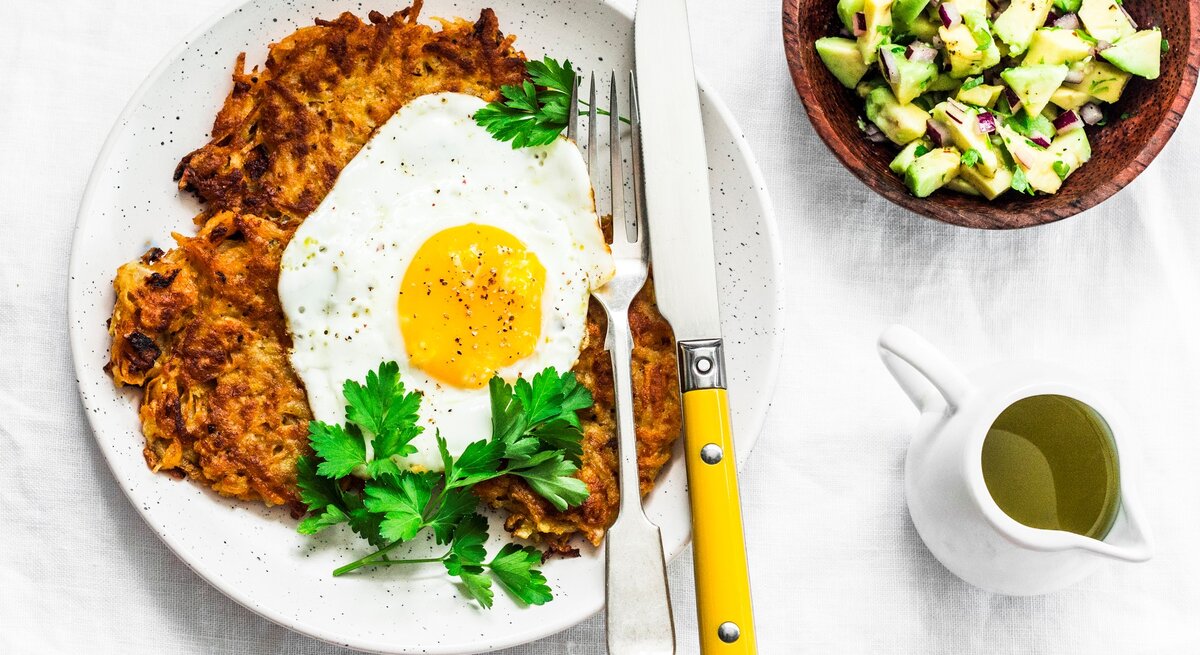 Ein weißer Teller mit den Röstis und Spiegelei auf einem weißen Tisch. Die Avocado steht daneben in einer kleinen braunen Schale.