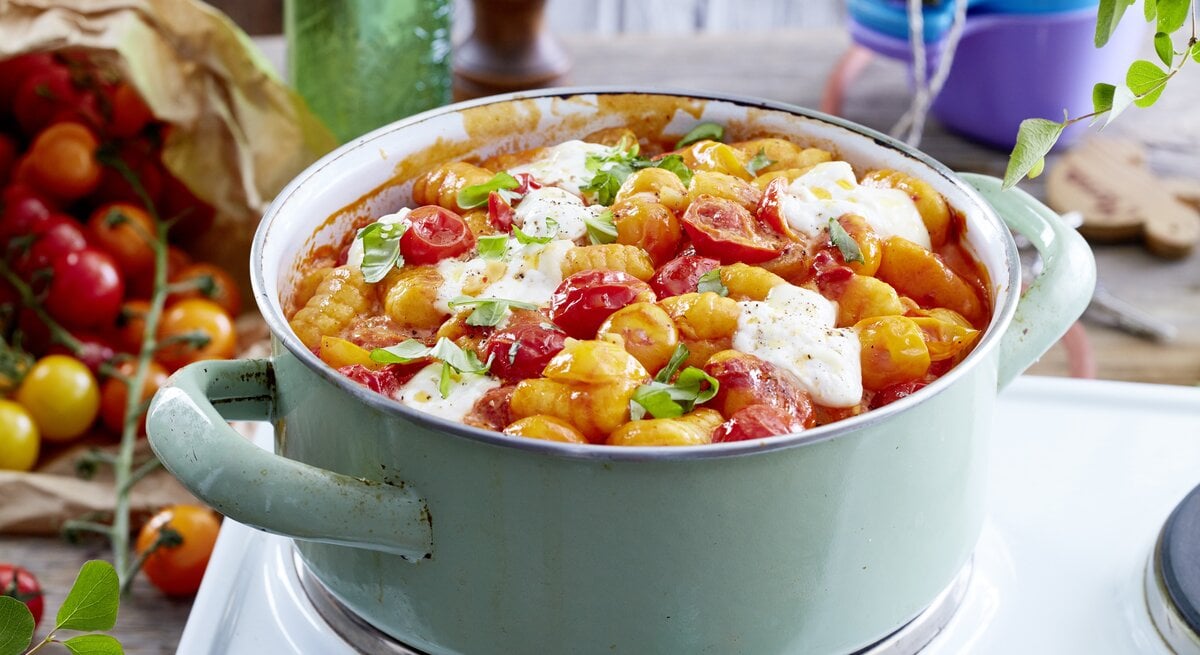 Eintopf aus Gnocchi mit Burrata und gebackenen Tomaten, bestreut mit frischem Basilikum und serviert in einem großen, hellen Kochtopf auf einem Campingtisch.  