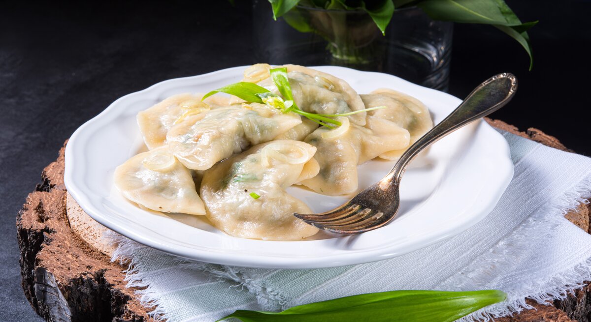 Frontal: Ein weißer Porzellanteller mit Bärlauch-Maultaschen steht auf einem weißen Tuch, das auf einem dunklen Untergrund liegt. Im Vorder- und Hintergrund sind frische Bärlauch-Blätter.