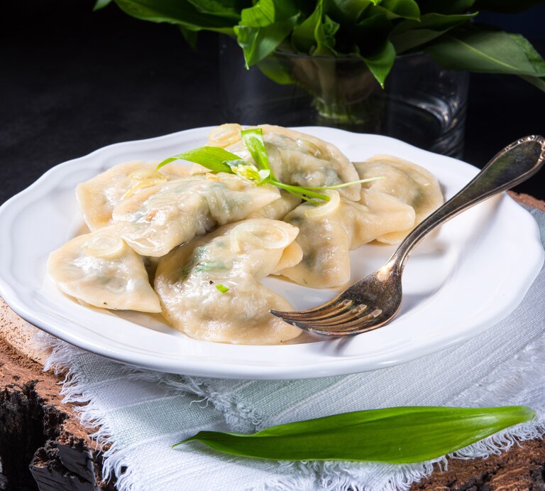 Frontal: Ein weißer Porzellanteller mit Bärlauch-Maultaschen steht auf einem weißen Tuch, das auf einem dunklen Untergrund liegt. Im Vorder- und Hintergrund sind frische Bärlauch-Blätter. | © Shutterstock / Dar1930