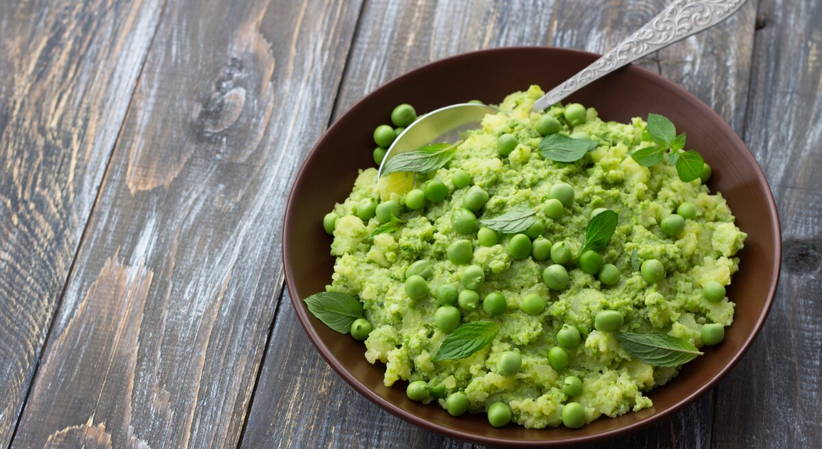 Draufsicht: Ein Teller mit Mushy Peas, also pürierten Erbsen, steht auf einen Holztisch. In dem Püree ist ein Löffel aus Metall.