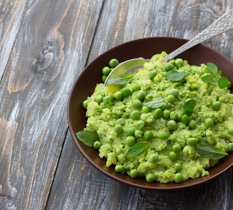 Draufsicht: Ein Teller mit Mushy Peas, also pürierten Erbsen, steht auf einen Holztisch. In dem Püree ist ein Löffel aus Metall. | © Shutterstock / olepeshkina