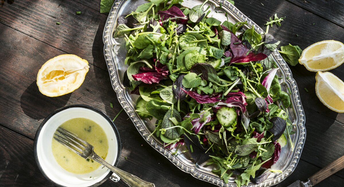 Silberne Platte mit Salat auf hölzernem Untergrund. Daneben einige ausgepresste Zitronen und eine Schale mit Buttermilch-Zitronen-Dressing. 