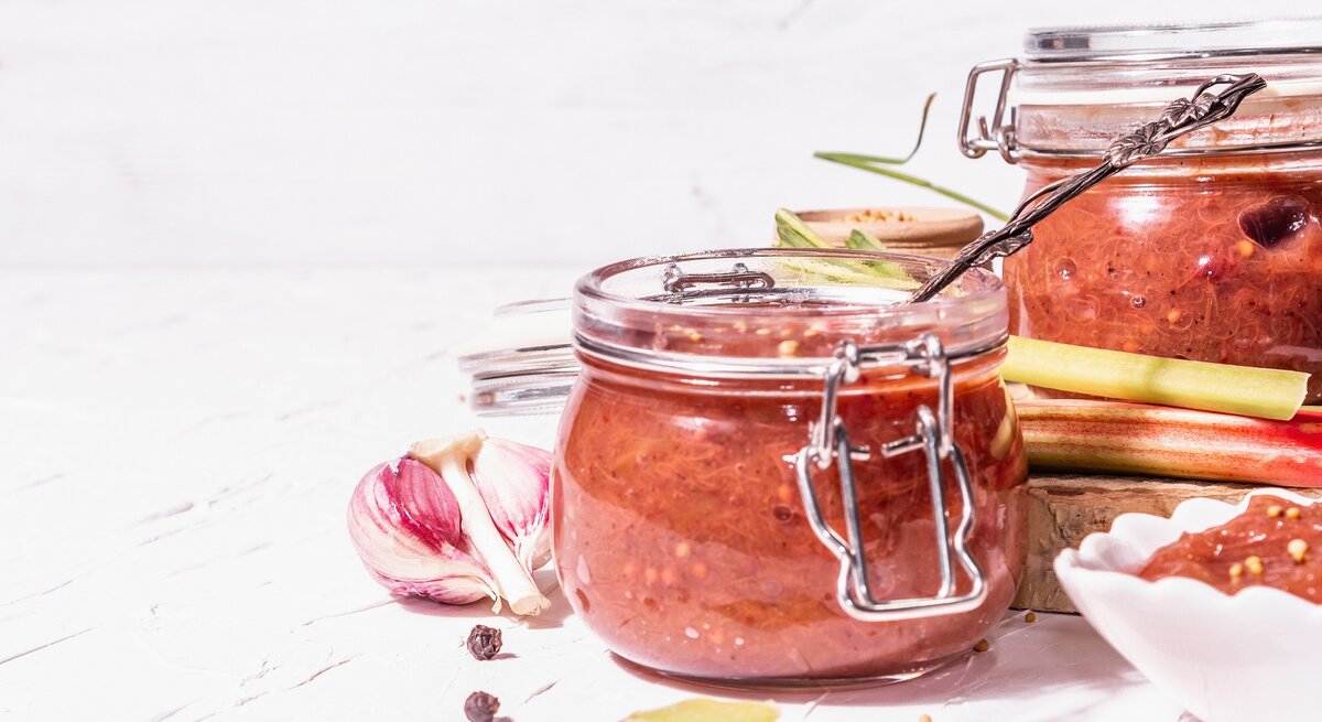 Zwei Weckgläser mit dem Rhabarber-Ketchup vor weißem Hintergrund und auf weißem Tisch. Drum herum einiges an Gewürzen wie Knoblauch oder Senfsaat.