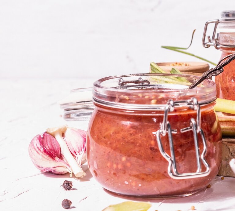 Zwei Weckgläser mit dem Rhabarber-Ketchup vor weißem Hintergrund und auf weißem Tisch. Drum herum einiges an Gewürzen wie Knoblauch oder Senfsaat. | © Shutterstock/ Fuzull Hanum