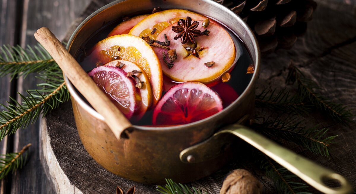 Ein Topf mit Glühwein, Orangenscheiben und Gewürzen steht auf einem Holzuntergrund, daneben liegen Tannengrün und Tannenzapfen.