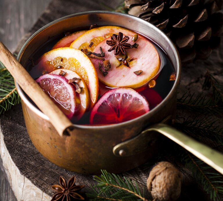 Ein Topf mit Glühwein, Orangenscheiben und Gewürzen steht auf einem Holzuntergrund, daneben liegen Tannengrün und Tannenzapfen. | © Getty Images/ Westend61