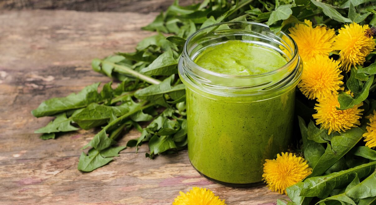 Ein Glas mit dem Löwenzahn-Pesto auf einem Holzuntergrund mit frischem Löwenzahn im Hintergrund.