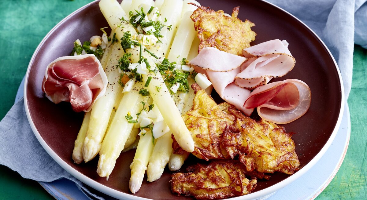 Auf einem Teller ist eine Portion Spargel mit Rösti und Schinken angerichtet. Sie ist mit Aufschnitt verfeinert.