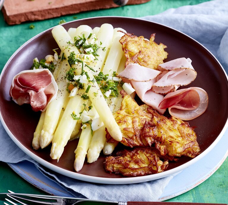 Auf einem Teller ist eine Portion Spargel mit Rösti und Schinken angerichtet. Sie ist mit Aufschnitt verfeinert. | © EAT CLUB/Isabell Triemer