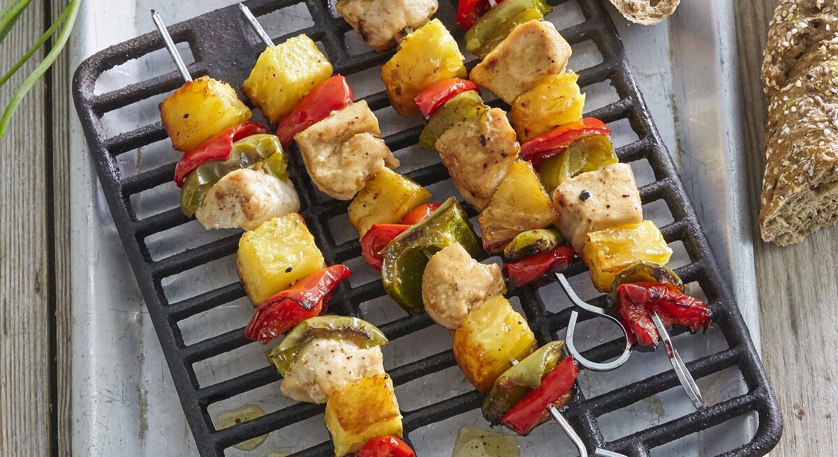 Mehrere Hawaii-Spieße mit Huhn und Ananas auf einem Grillgitter, daneben Brot in der Draufsicht.