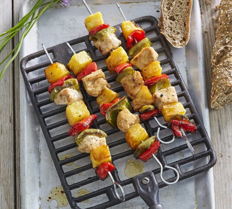 Mehrere Hawaii-Spieße mit Huhn und Ananas auf einem Grillgitter, daneben Brot in der Draufsicht. | © StockFood / Profimedia