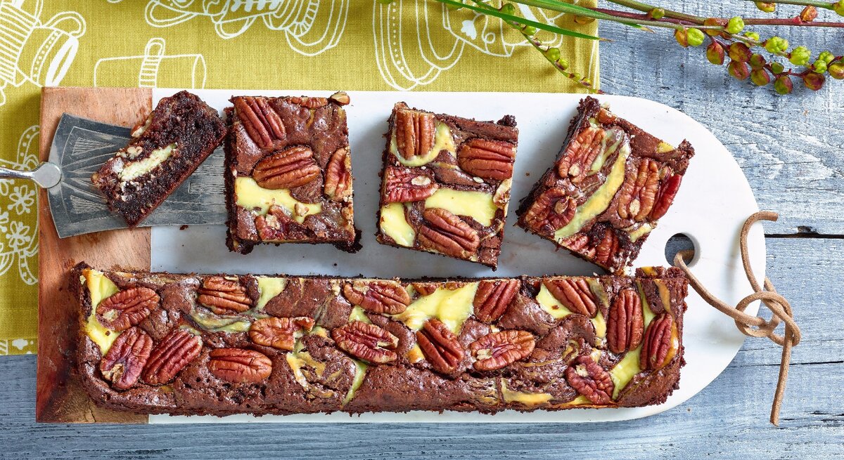 Brownies mit Frischkäse und Pekannuss auf weißem Schneidebrett, gelb-grünem Tuch und blauem Tisch. Brownies der Länge nach halbiert. Eine Hälfte zu Rechtecken geschnitten. Das Brett ist gelocht und eine Lederschnur durchgefädelt. Links ein Stück Brwonie auf silbernen Tortenheber mit Intarsien. Oben Zweige.