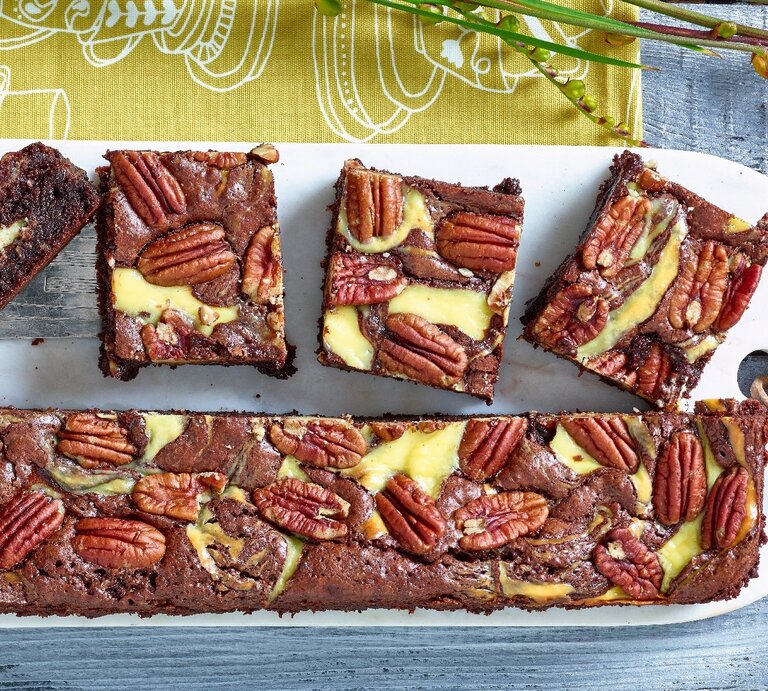 Brownies mit Frischkäse und Pekannuss auf weißem Schneidebrett, gelb-grünem Tuch und blauem Tisch. Brownies der Länge nach halbiert. Eine Hälfte zu Rechtecken geschnitten. Das Brett ist gelocht und eine Lederschnur durchgefädelt. Links ein Stück Brwonie auf silbernen Tortenheber mit Intarsien. Oben Zweige. | © Matthias Liebich