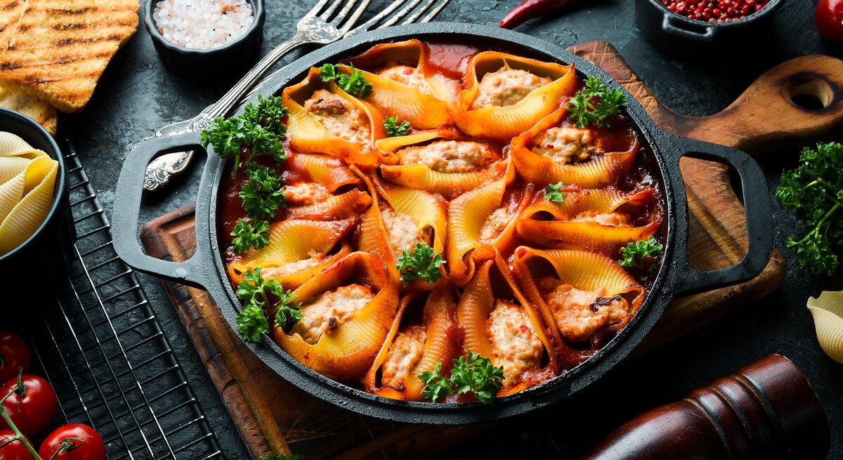 Draufsicht: eine dunkle Pfanne mit gefüllten Muschelnudeln mit Schmand-Hackfleisch steht auf einem Holzbrettchen. Drumherin liegen Schälchen mit Gewürzen, eine pfeffermüfle, Tomaten und rohen Muschelnudeln.