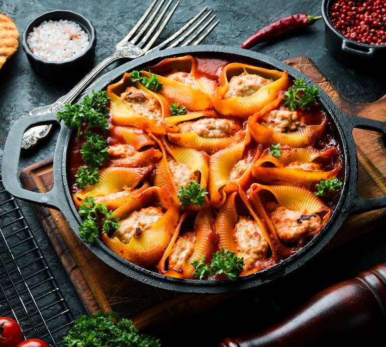 Draufsicht: eine dunkle Pfanne mit gefüllten Muschelnudeln mit Schmand-Hackfleisch steht auf einem Holzbrettchen. Drumherin liegen Schälchen mit Gewürzen, eine pfeffermüfle, Tomaten und rohen Muschelnudeln. | © Shutterstock / YARUNIV Studio