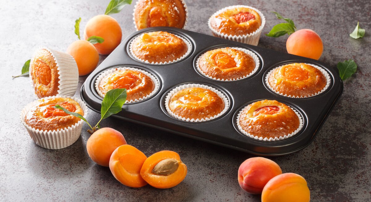 Muffingblech mit Aprikosen-Joghurt-Muffins. Daneben liegen einzelne Muffins und Aprikosen. 