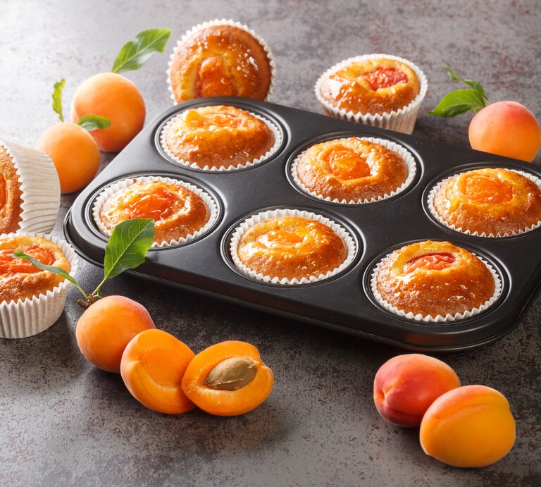 Muffingblech mit Aprikosen-Joghurt-Muffins. Daneben liegen einzelne Muffins und Aprikosen.  | © Getty Images/ALLEKO
